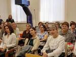 В зале на мероприятии группа женщин в деловой одежде сидит в креслах, на фоне — окна с вертикальными жалюзи, над аудиторией — мультимедийный проектор.