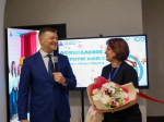 На фото улыбающийся мужчина в тёмно‑синем костюме с микрофоном беседует с женщиной в чёрном наряде, держащей букет цветов; на фоне — экран с надписью об официальном открытии кабинета дошкольного образования и символикой Года дошкольного образования.