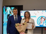 На фото мужчина в тёмно‑синем костюме стоит рядом с улыбающейся женщиной в светлом пиджаке, которая держит в руках букет цветов и грамоту; на фоне — экран с символикой «Года дошкольного образования».