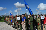 На фото — строй участников военно‑патриотического мероприятия: молодые люди в военной форме и гражданской одежде держат флаги и таблички с названиями районов, на фоне — ясное небо с облаками.