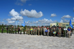 На фото — строй участников военно‑патриотического мероприятия на площади у спортивного комплекса: люди в военной форме и гражданской одежде с флагами и табличками, на фоне — голубое небо с облаками.