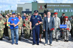 На фото — группа людей (в том числе военнослужащие, гражданские лица и школьники) с красными гвоздиками участвует в памятном мероприятии у спортивного комплекса, в центре — мужчина в форменной одежде и мужчина в деловом костюме.