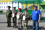 На фото — офицер в военной форме и мужчина в синей футболке стоят рядом с четырьмя девушками в камуфляжной форме, держащими грамоты и медалями, на фоне здания и зелёной навеса.