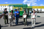 На фото — церемония награждения участников военно‑патриотических сборов: девушки в камуфляжной форме с грамотами и медалями стоят на фоне современного здания и зелёной конструкции, рядом — военнослужащие и организаторы мероприятия.