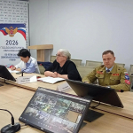 В конференц-зале за столом сидят участники (в том числе сотрудник в форменной одежде), перед ними — таблички с именами, документы и мониторы, на одном из экранов — окно видеоконференции.