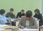 Группа женщин в деловой обстановке работает за столами с документами, блокнотами, ручками и канцелярскими принадлежностями в светлом помещении с однотонной стеной.