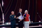 На сцене во время торжественной церемонии мужчина в деловом костюме вручает женщине в красно‑белом наряде награду, рядом стоит военный в форме и жестом сопровождает момент награждения, на фоне — стилизованный декор и сценическое освещение.