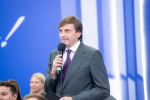 Министр просвещения РФ Сергей Кравцов в сером костюме и фиолетовом галстуке выступает с микрофоном в руке на фоне сине‑белого оформления, позади него — слушатели.