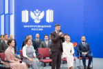 На фото — мужчина в костюме с микрофоном выступает перед аудиторией на фоне стены с эмблемой и надписью «Минпросвещения России», позади него слушатели в деловой одежде сидят на стульях.