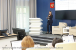 Эксперт ФИЦТО Татьяна Покидышева в деловом костюме выступает с микрофоном у зигзагообразной трибуны в конференц‑зале перед аудиторией; на фоне — герб РФ и экран с презентацией Минпросвещения России и Рособрнадзора.